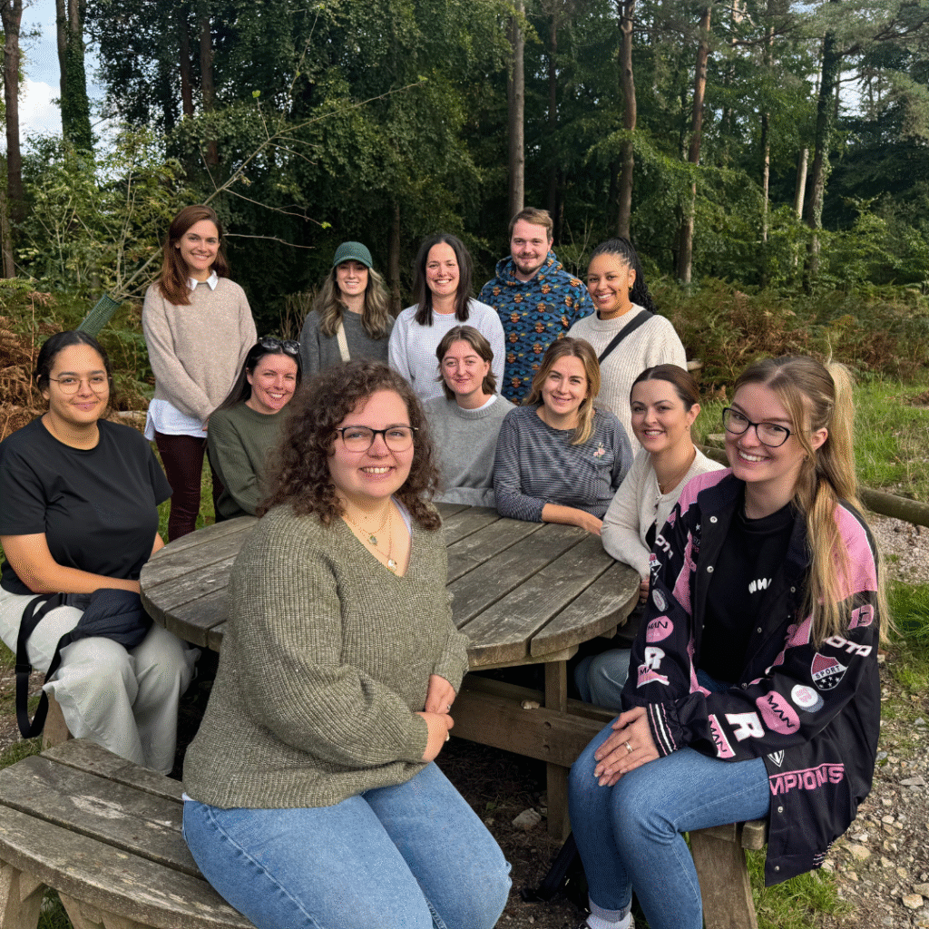 A group of trainee mental health practitioners sit around a wooden bench in the woods.