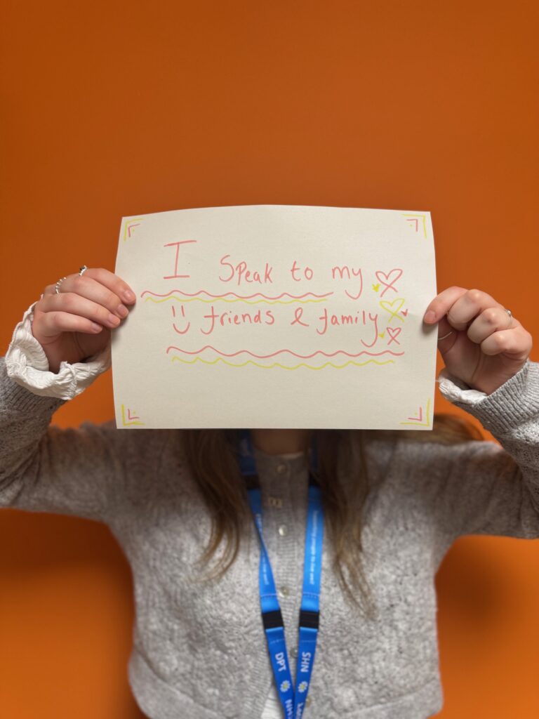 A person infront of an orange wall holding up a peice of A4 paper with the words 'I speak to my friends and family'