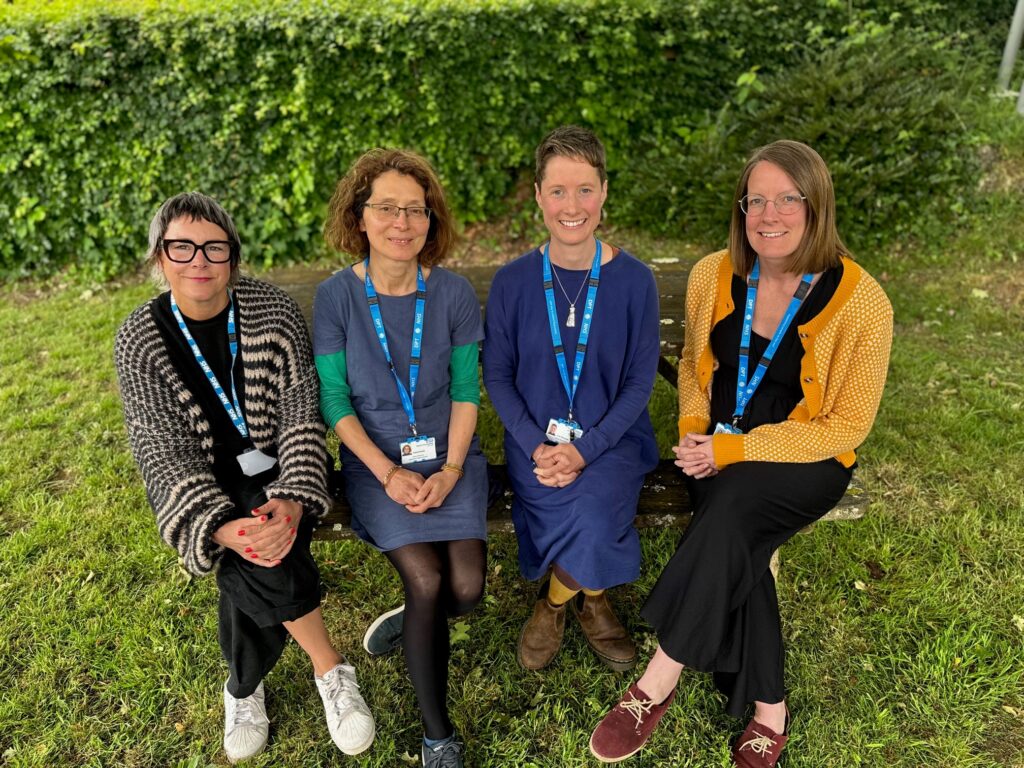 Four members of the PAIRs team sit on a bench, smiling at the camera. All women are wearing CFHD lanyards
